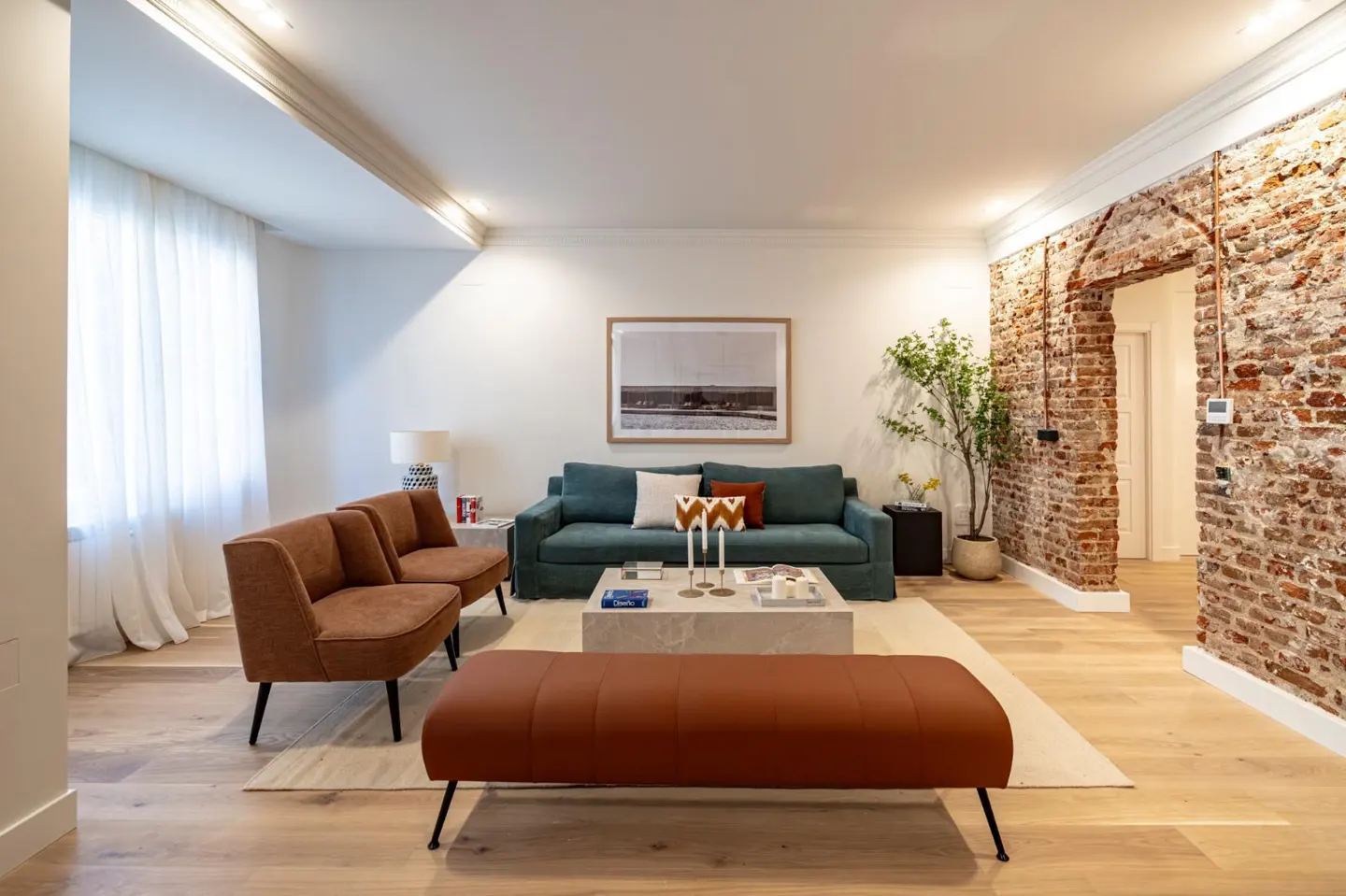 Bright living room with a teal sofa, brown chairs, and a bench on a light wood floor. A brick wall with an open doorway adds texture.