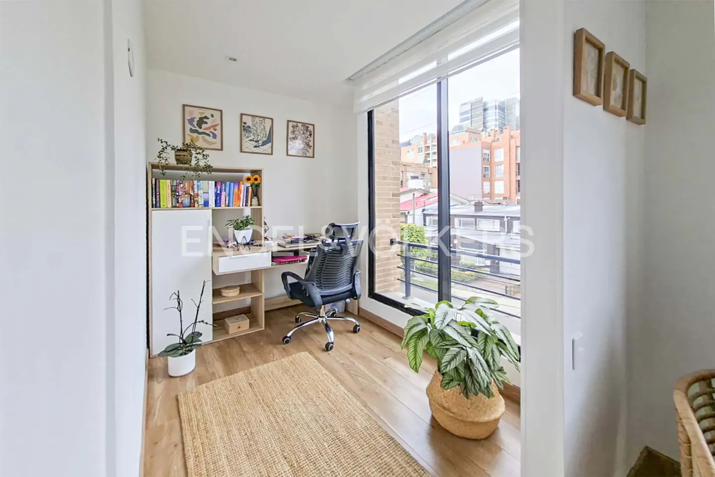 Bright home office with a desk, chair, and bookshelf. A large window offers a city view. A jute rug and potted plants add warmth.
