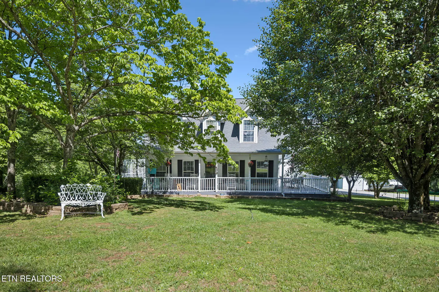 A white house with a porch and black shutters sits behind a green lawn and trees. A white bench is on the left.