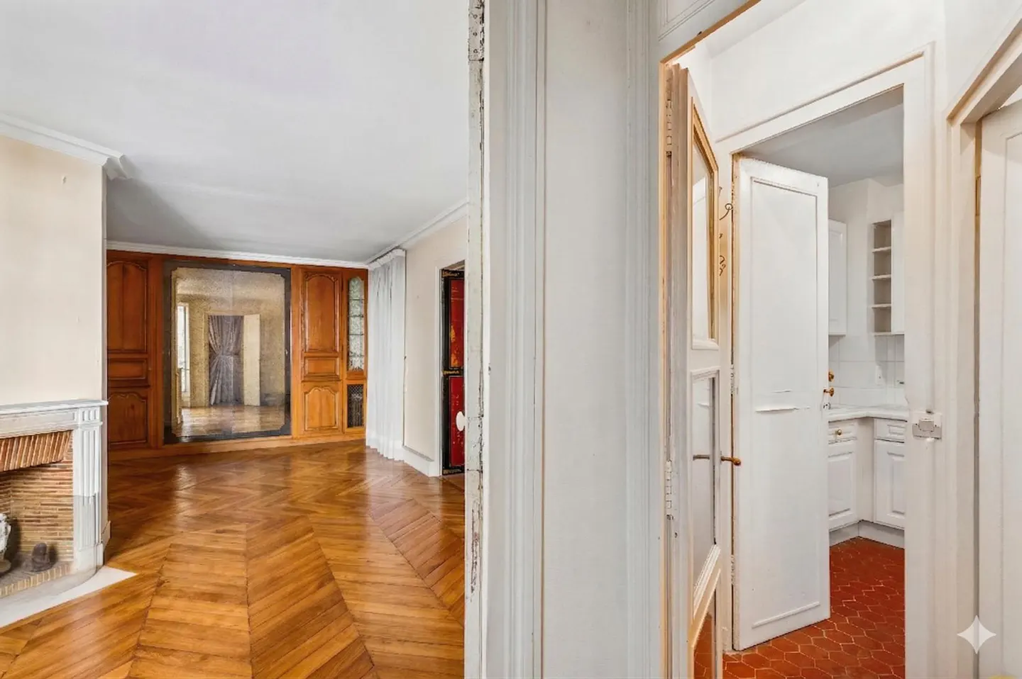 Interior view of a home with herringbone wood floors, a fireplace, and an open doorway to a white kitchen.