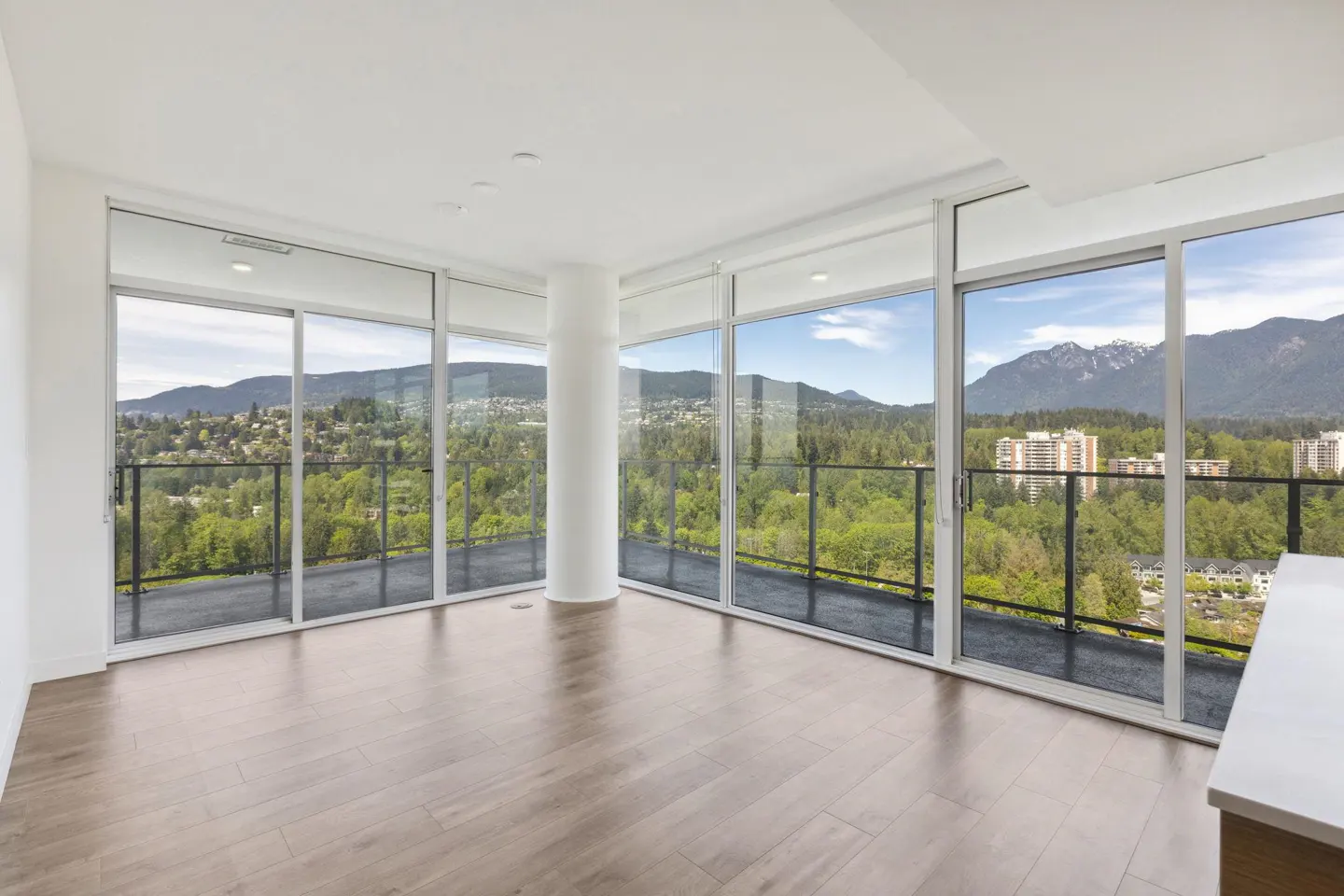 Bright, empty room with wood floors and floor-to-ceiling windows. A balcony overlooks a green landscape with mountains in the distance.