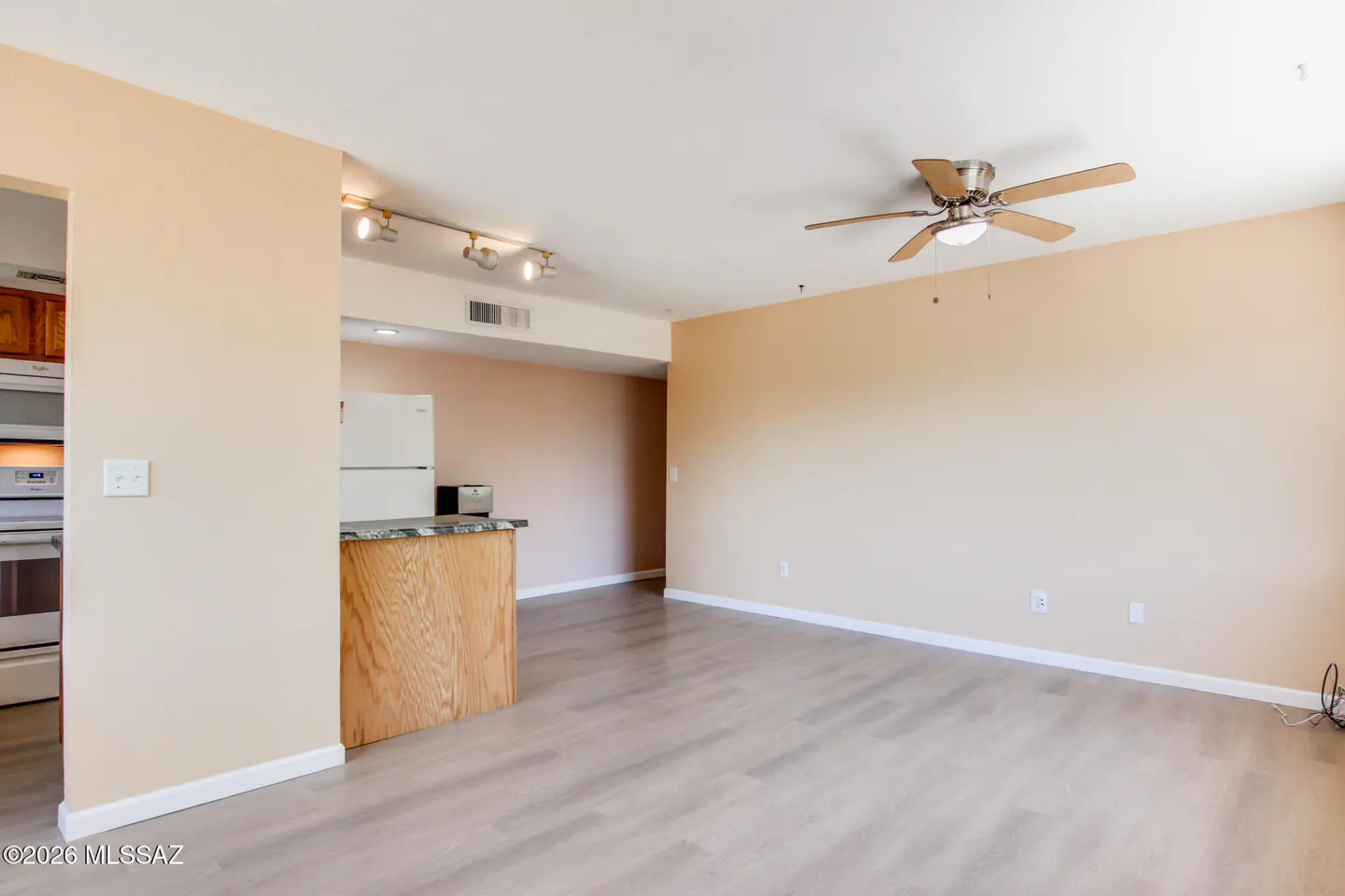A bright, empty living room with beige walls, light wood floors, and a ceiling fan. A kitchen area is visible in the background.