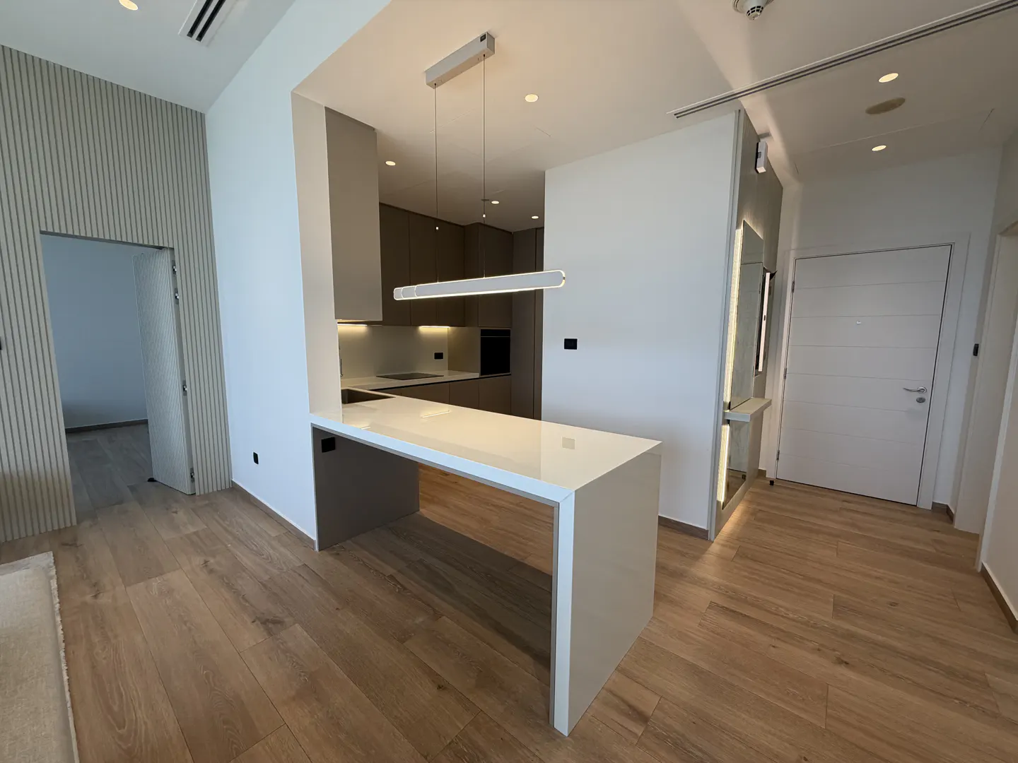 A modern apartment interior with wood floors, a white kitchen island, and a linear pendant light. A doorway and a white door are visible.