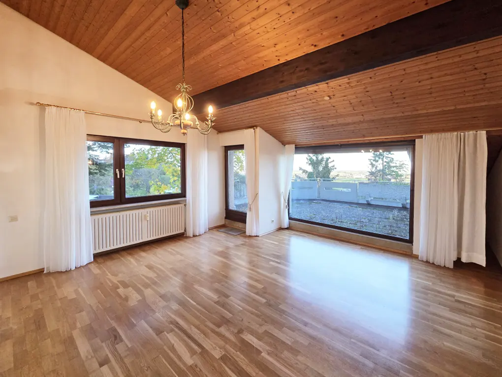 Bright room with wood floors, wood ceiling, and white walls. Large windows with white curtains offer outdoor views. A chandelier hangs from the ceiling.