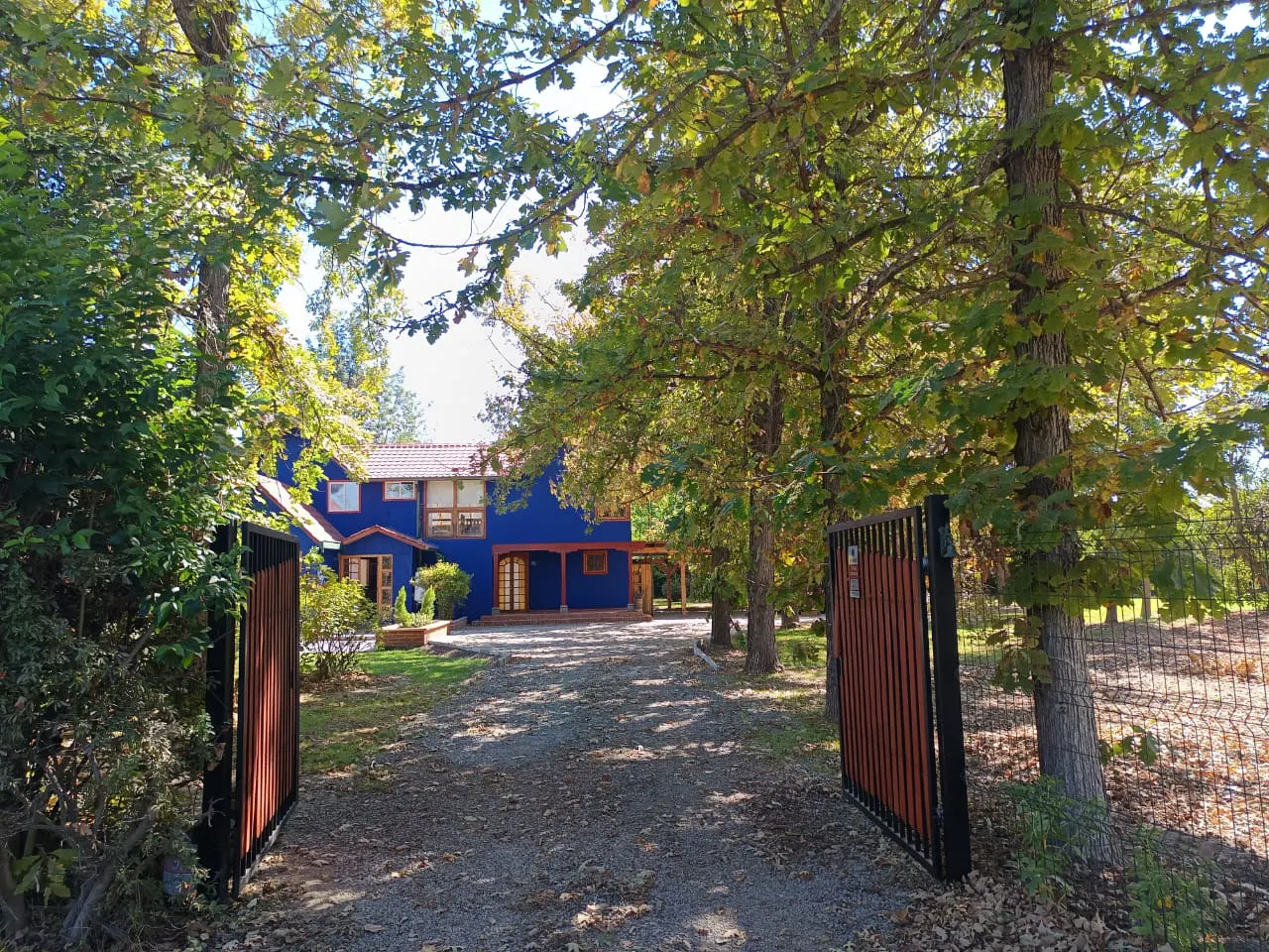 View through open gate to a two-story blue house with a red tile roof and gravel driveway. Trees surround the property.