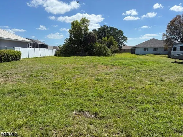 Vacant lot with green grass, trees, and houses in the background under a blue sky with white clouds. A white fence is on the left.