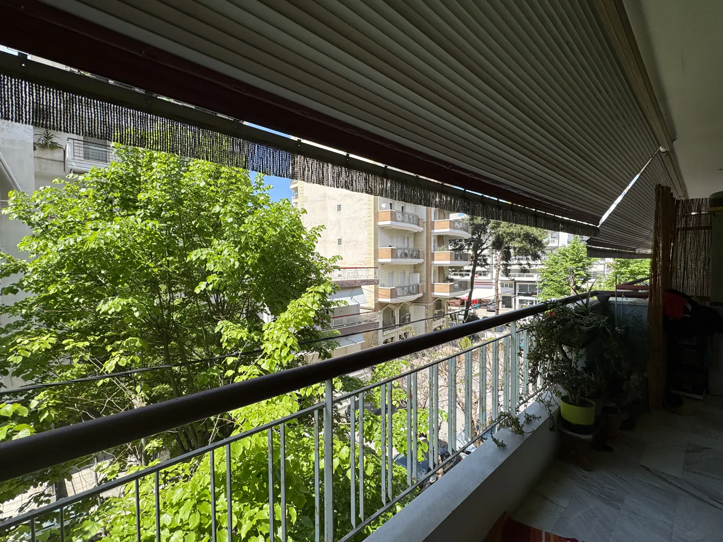 View from a balcony with a metal railing, overlooking green trees and buildings on a sunny day. A retractable awning provides shade.