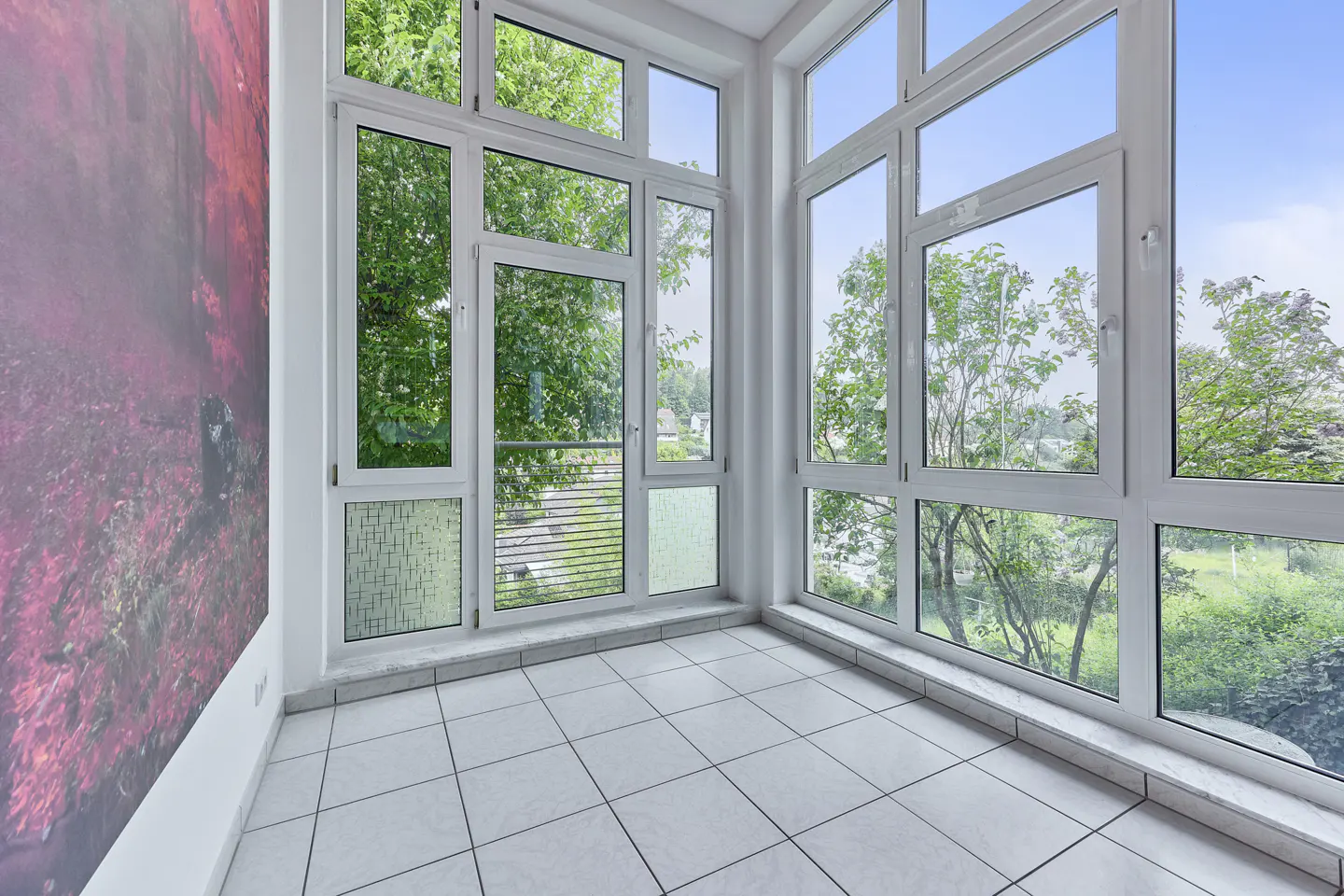 Bright sunroom with white tile floor and large white-framed windows showing green trees and blue sky. A red and gray abstract painting is on the left wall.