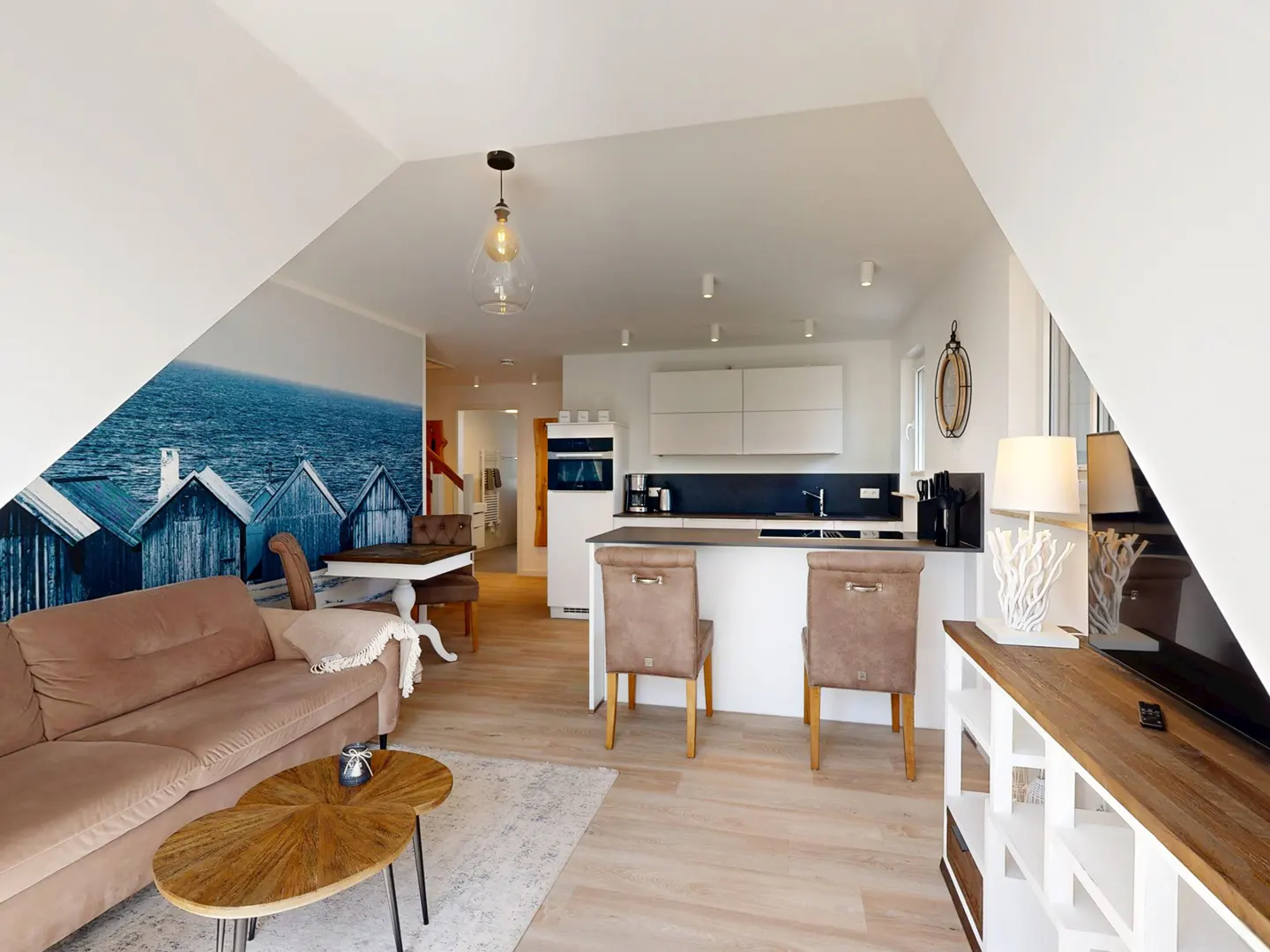 Modern open-plan living area with a beige sofa, wooden coffee tables, kitchen island with chairs, and a coastal wall mural.
