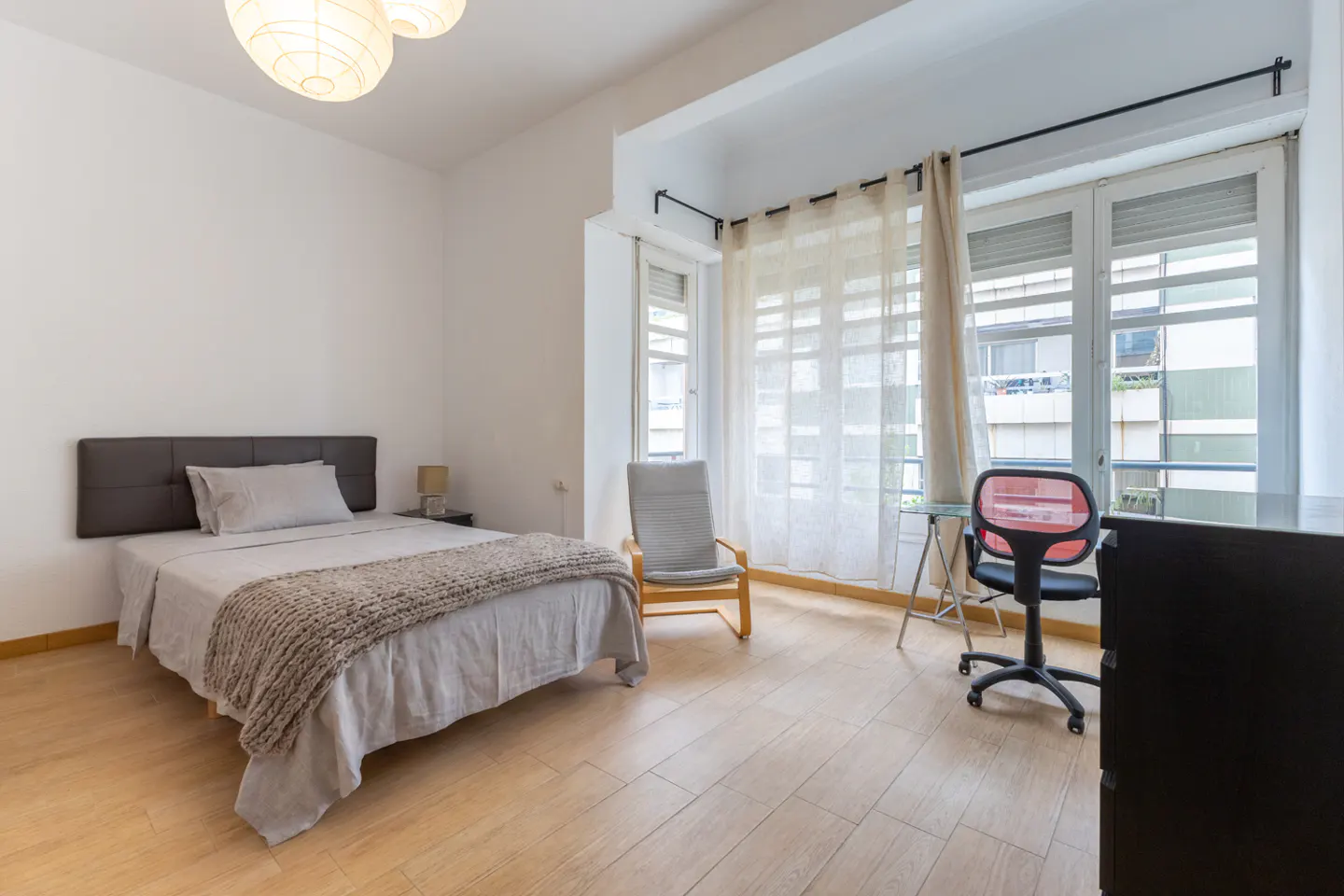 Bright bedroom with a bed, chair, desk, and large windows with sheer curtains. The walls are white and the floor is light wood.