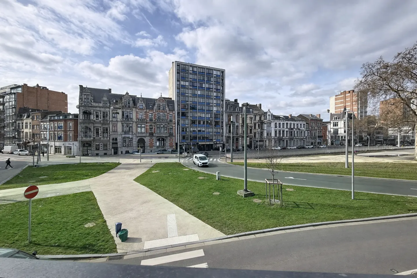 Cityscape view with a mix of old and modern buildings under a cloudy sky. A white van drives on the road, and green spaces are visible.