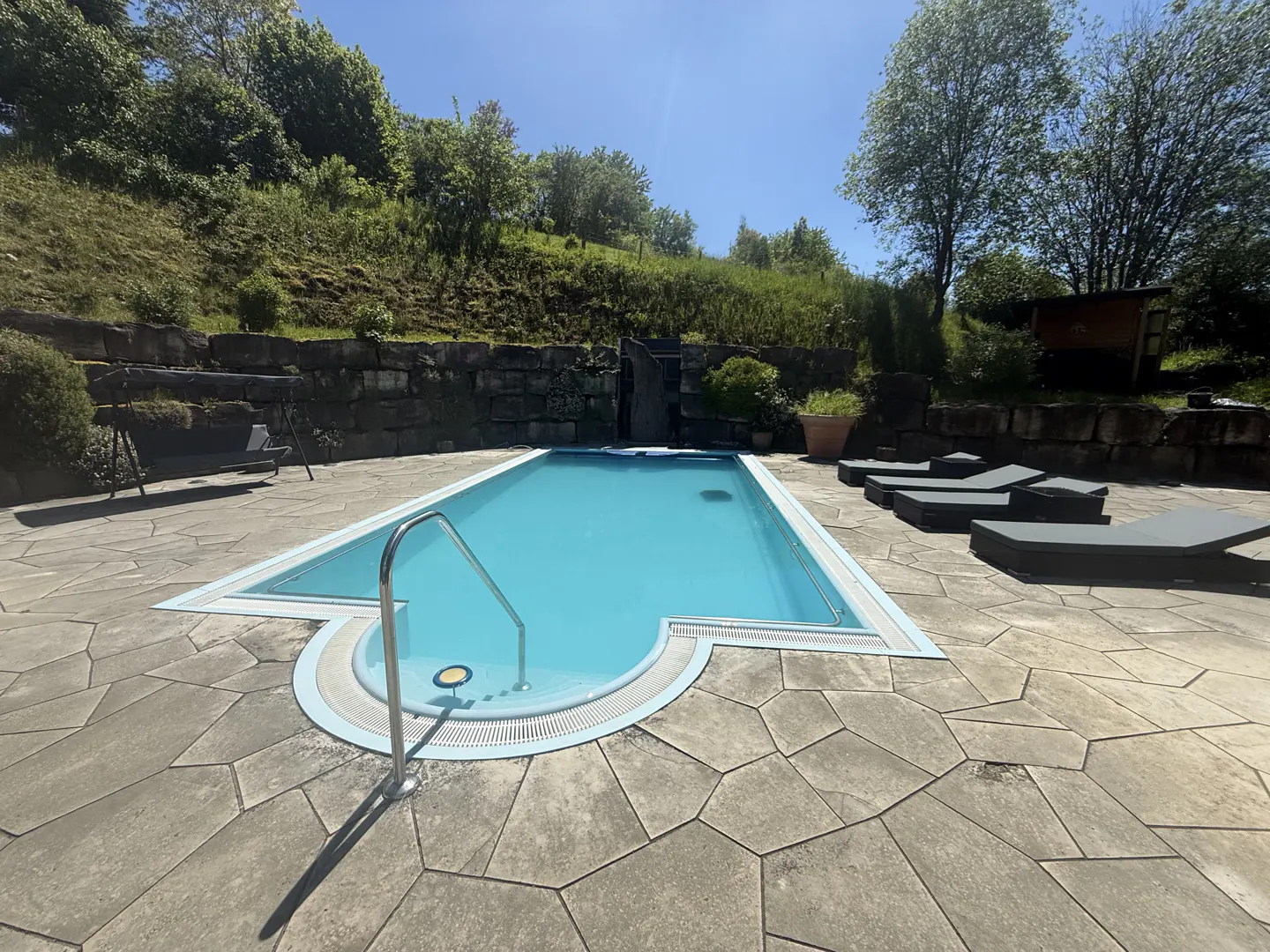 Outdoor pool with blue water, surrounded by stone patio, lounge chairs, and a green hillside.