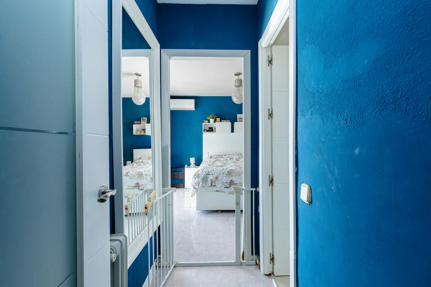 Hallway with blue walls and white trim leads to a bedroom with a bed and white furniture. A safety gate is installed in the doorway.