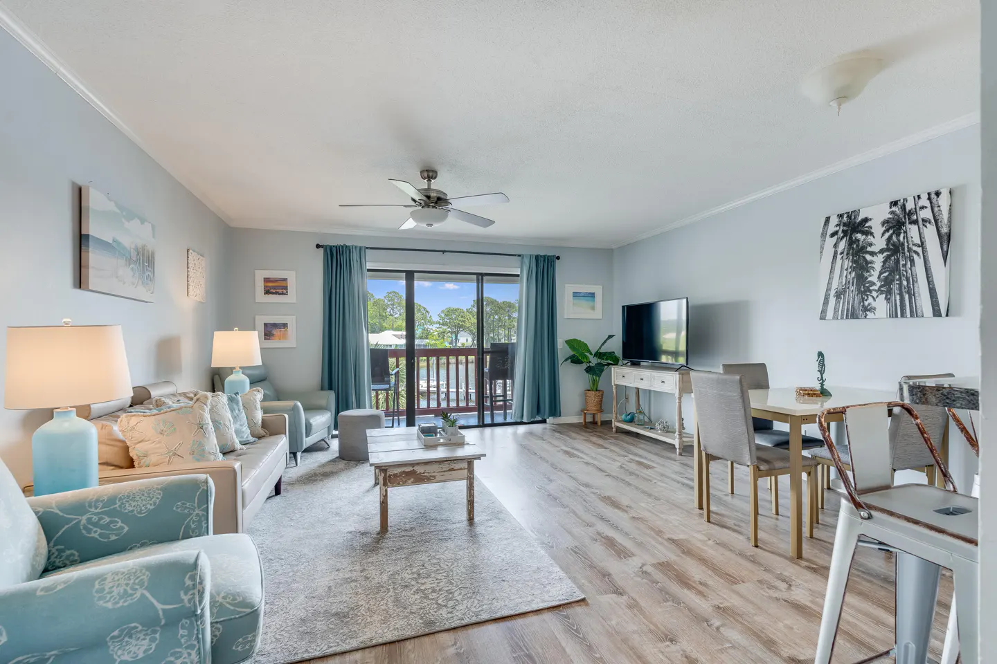 Living room with light blue walls, wood floors, and a sliding glass door to a balcony. Furnished with sofas, chairs, and a dining table.