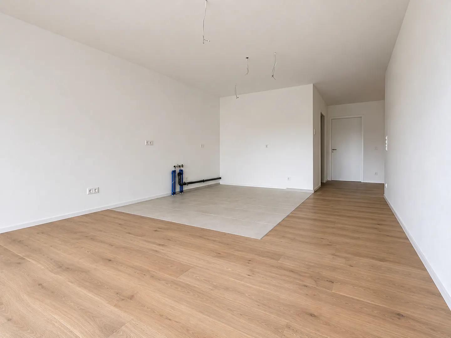 Empty room with light wood floors, white walls, and a section of gray tile. Blue pipes are visible on the wall.