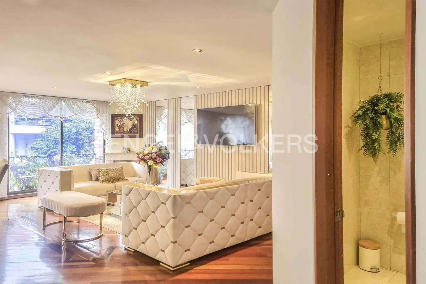 Living room with beige sofas, wood floors, and a chandelier. A bathroom is visible through a doorway with a hanging plant.