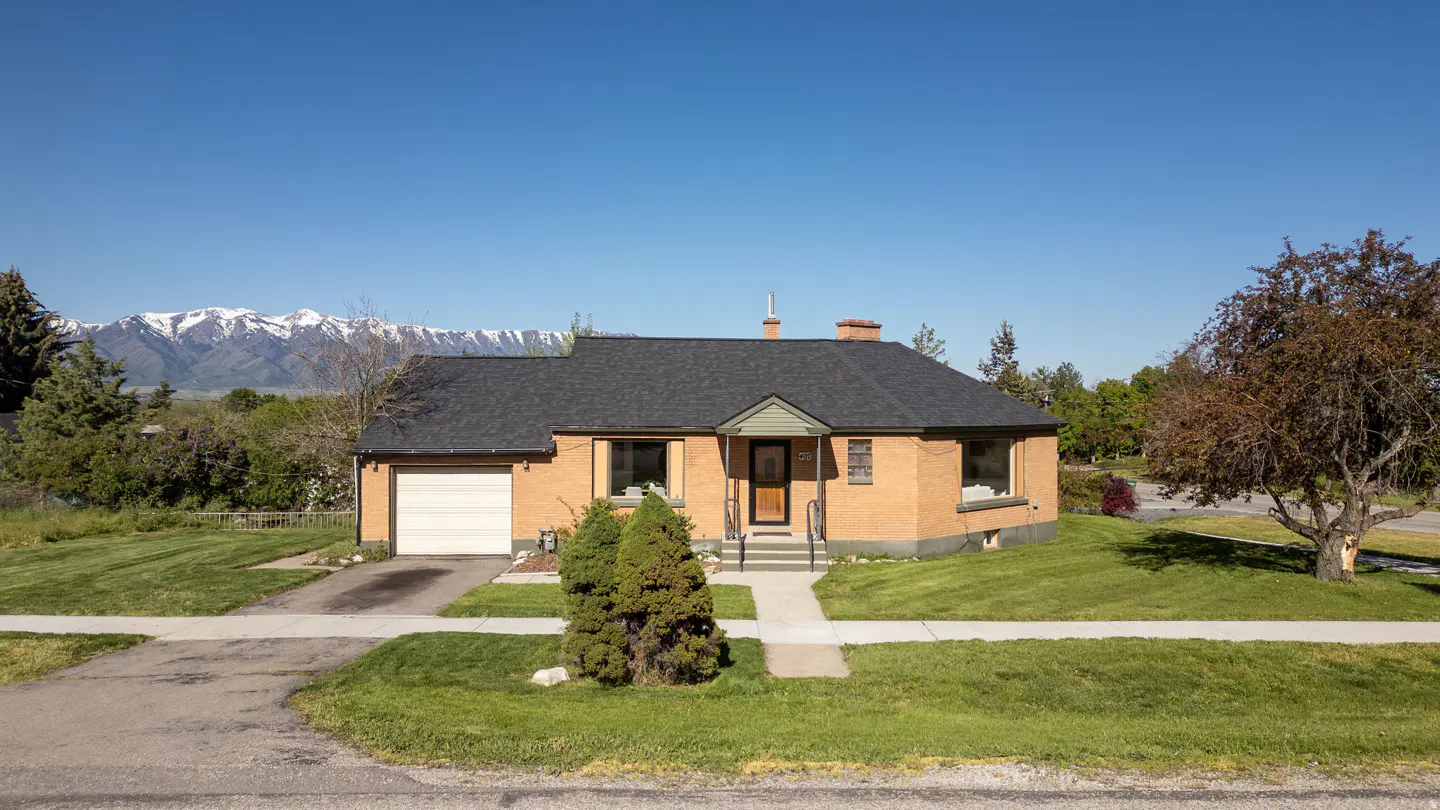 A single-story brick house with a dark roof, green lawn, and snow-capped mountains in the background.
