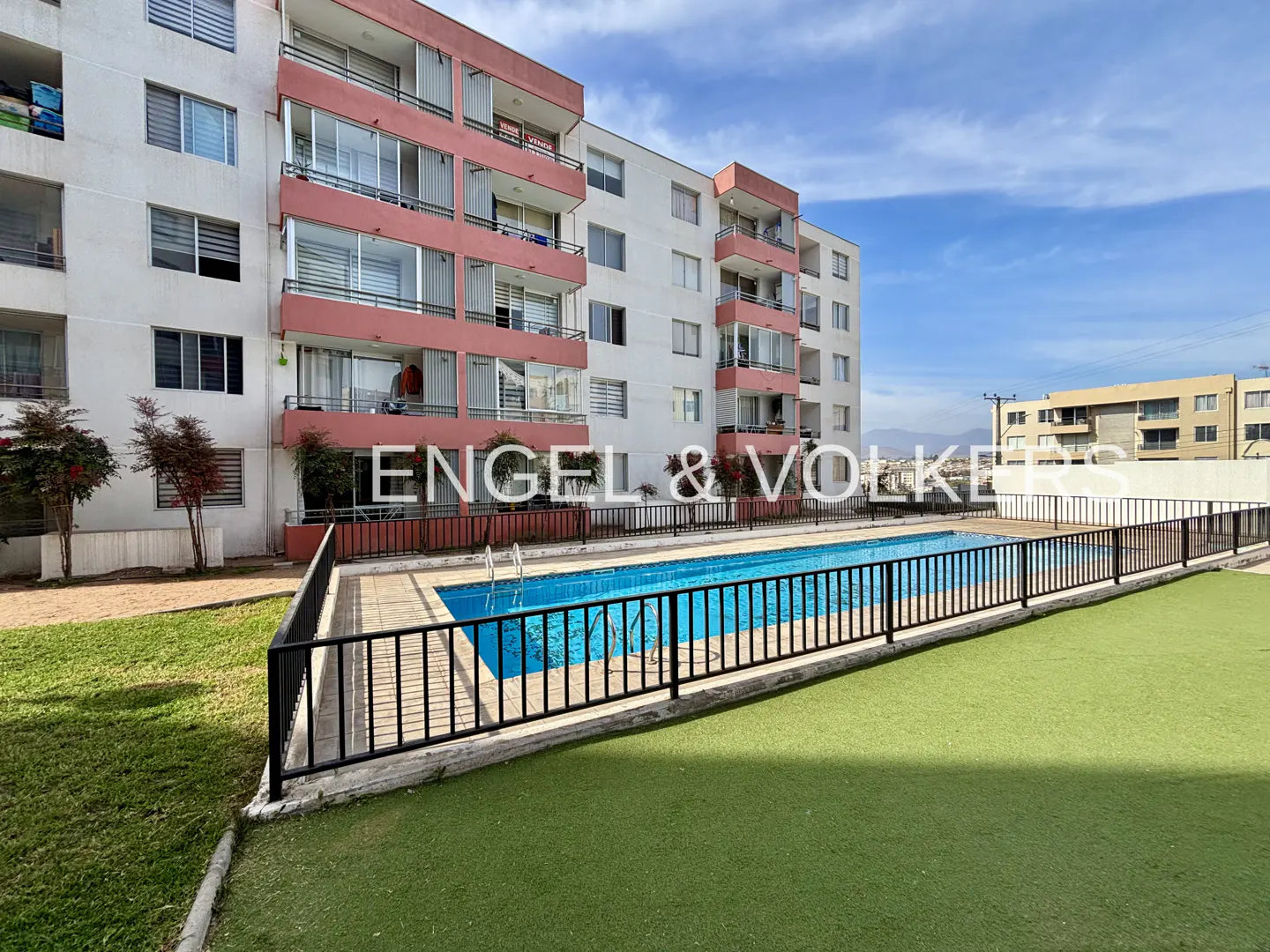 Exterior view of a white apartment building with pink balconies, a pool, and green turf.