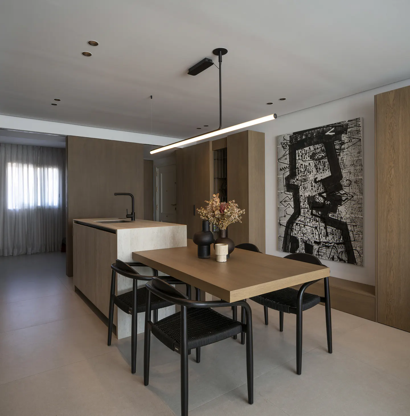Modern dining area with a wooden table, black chairs, a linear pendant light, and abstract art on a light-toned wall.