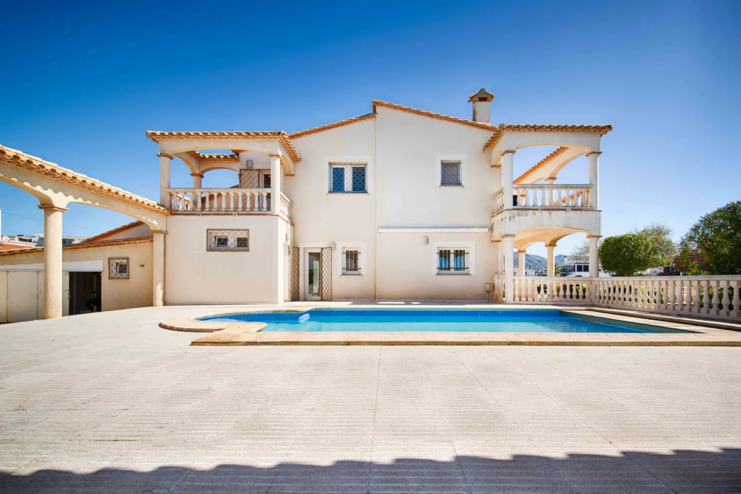 Exterior of a two-story beige house with a blue pool and balconies under a clear blue sky.
