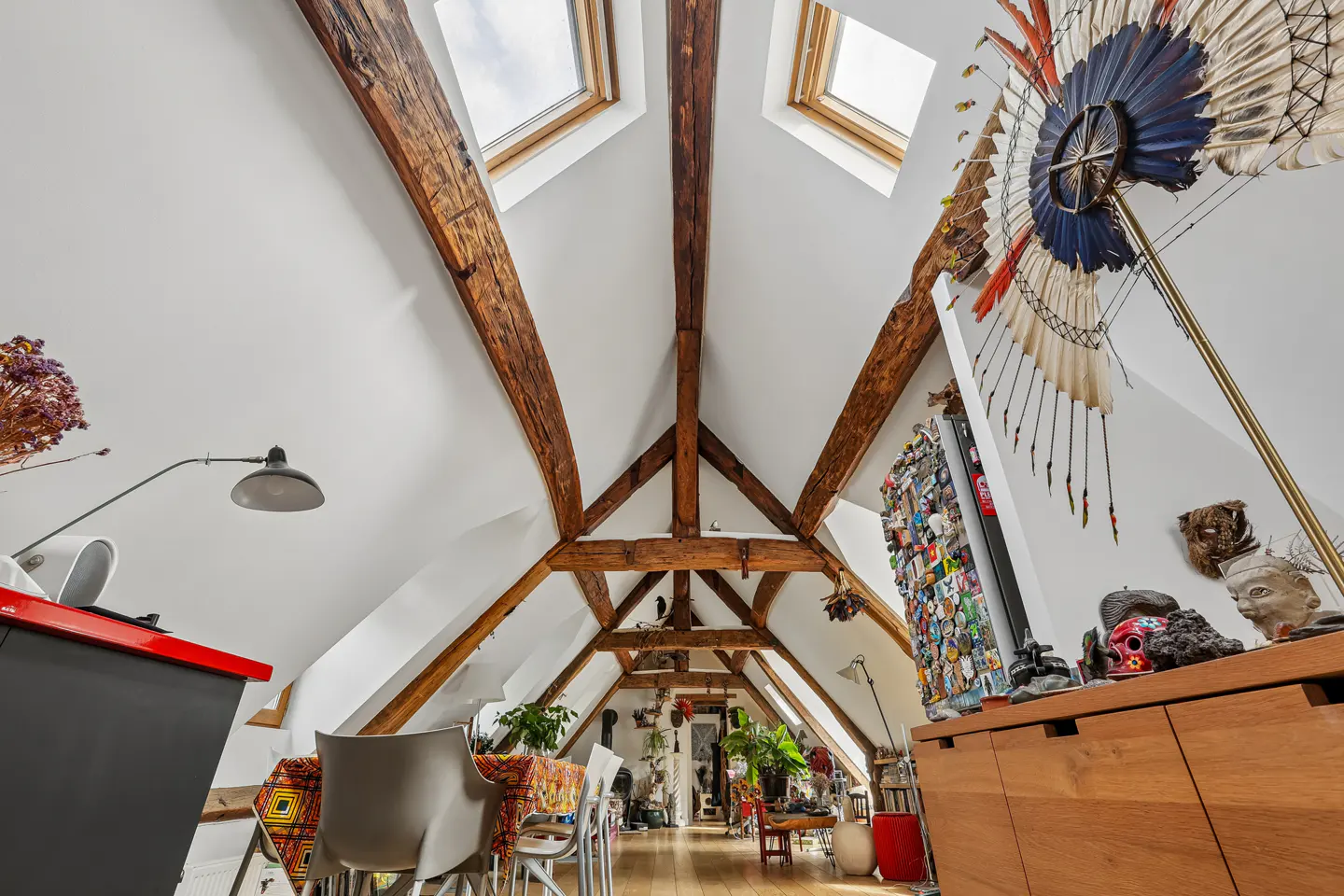 Attic room with exposed wooden beams, skylights, and eclectic decor. A dining table and chairs are visible, along with art and plants.
