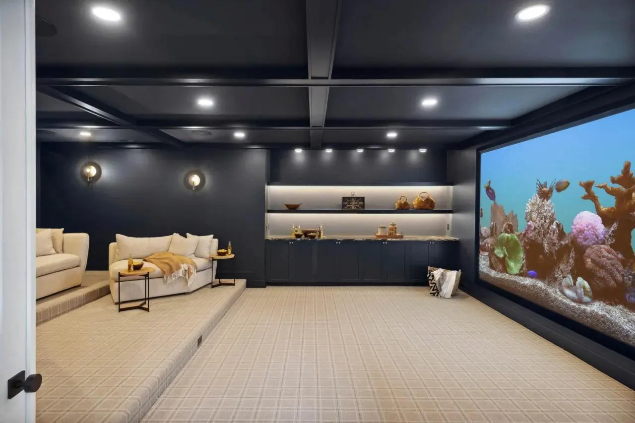 A home theater room with beige carpet, dark blue walls, and a large screen showing an aquarium scene. Two sofas sit on a raised platform.