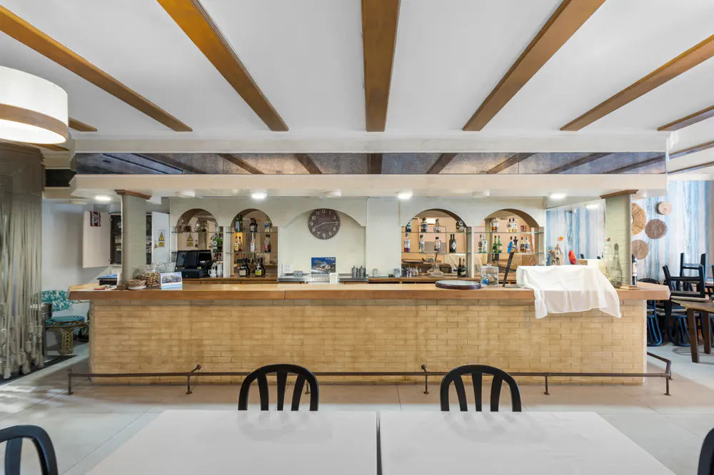 Interior view of a bar area with a brick counter, arched shelves displaying bottles, and tables with black chairs.