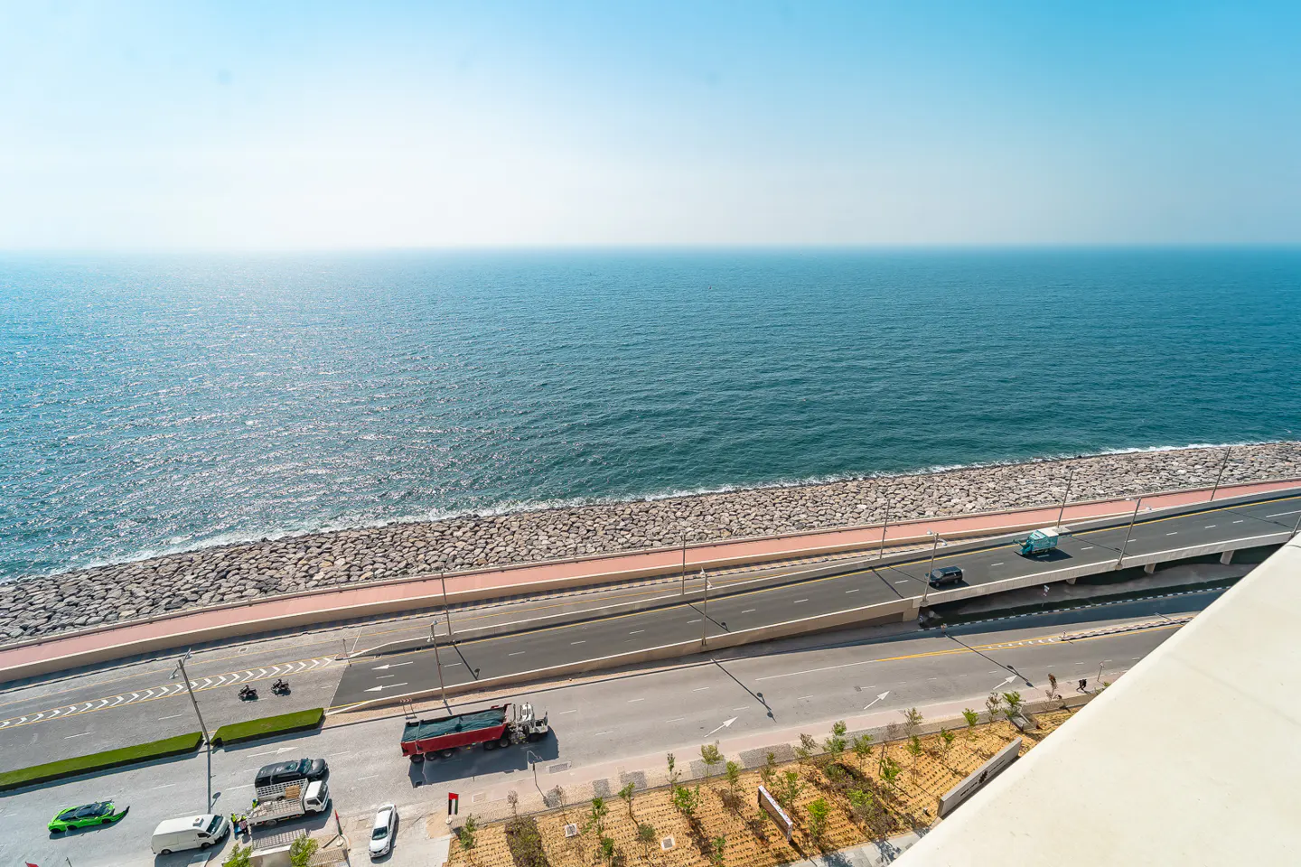 Aerial view of a coastal road with cars, a red truck, and a green sports car, next to a blue ocean under a clear sky.