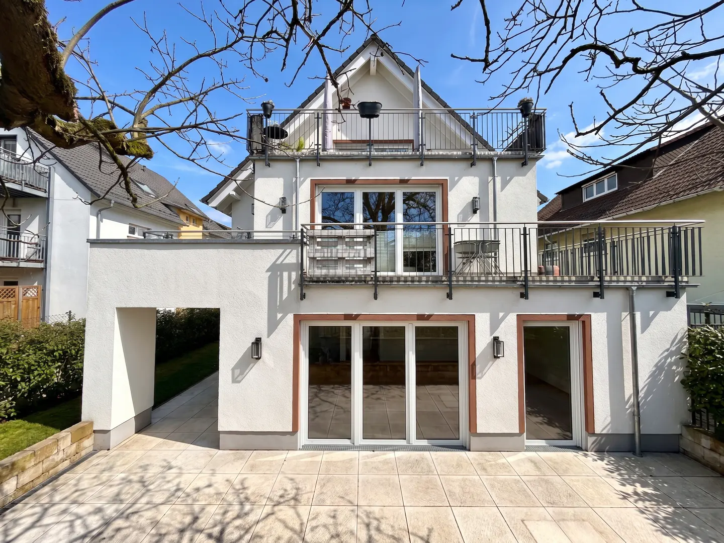 Two-story white house with a balcony and a patio on a sunny day. The house has large windows and a modern design.