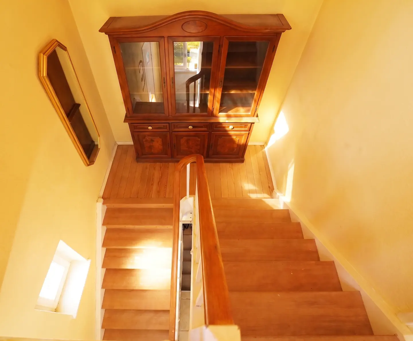 Wooden stairs lead to a landing with a dark wood china cabinet. A gold-framed mirror hangs on the yellow wall.