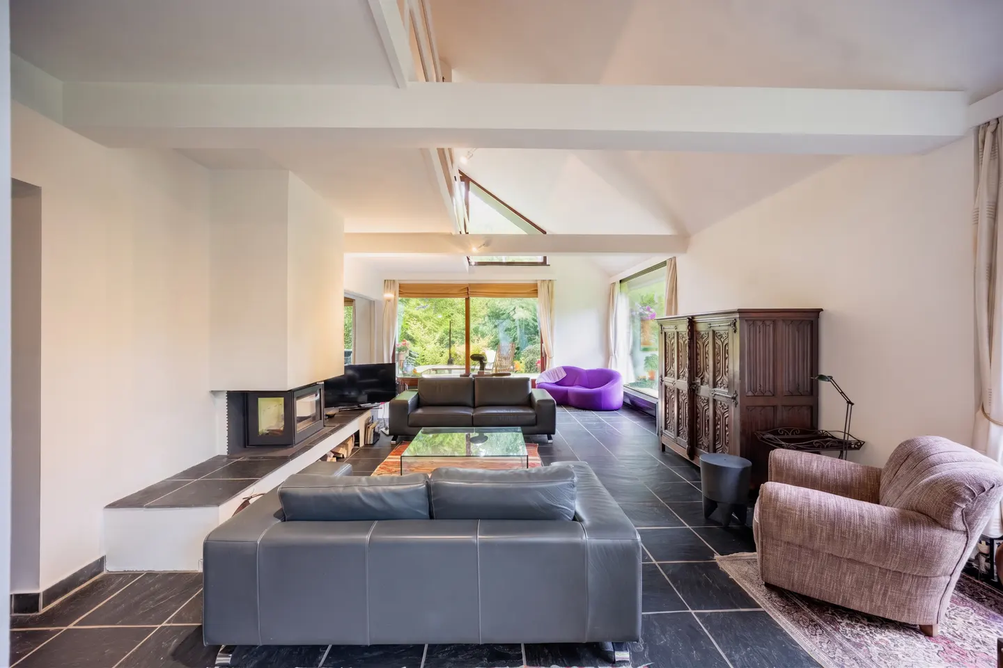 A bright living room with black tile floors, a gray leather sofa, and a fireplace. Large windows overlook a green yard.