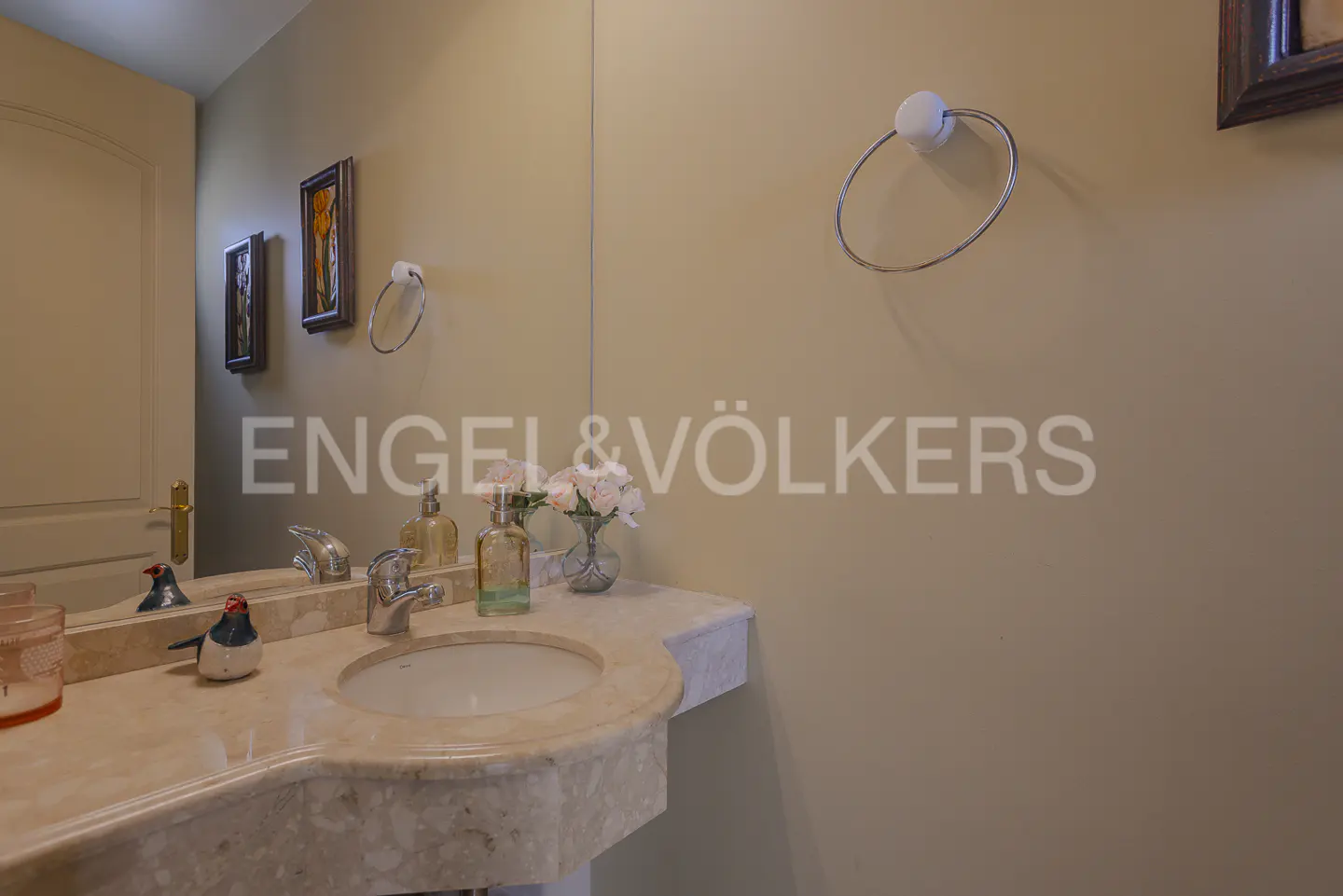 Bathroom with marble sink, silver faucet, and decorative bird. Beige walls with towel ring and framed art. Engel & Völkers logo.