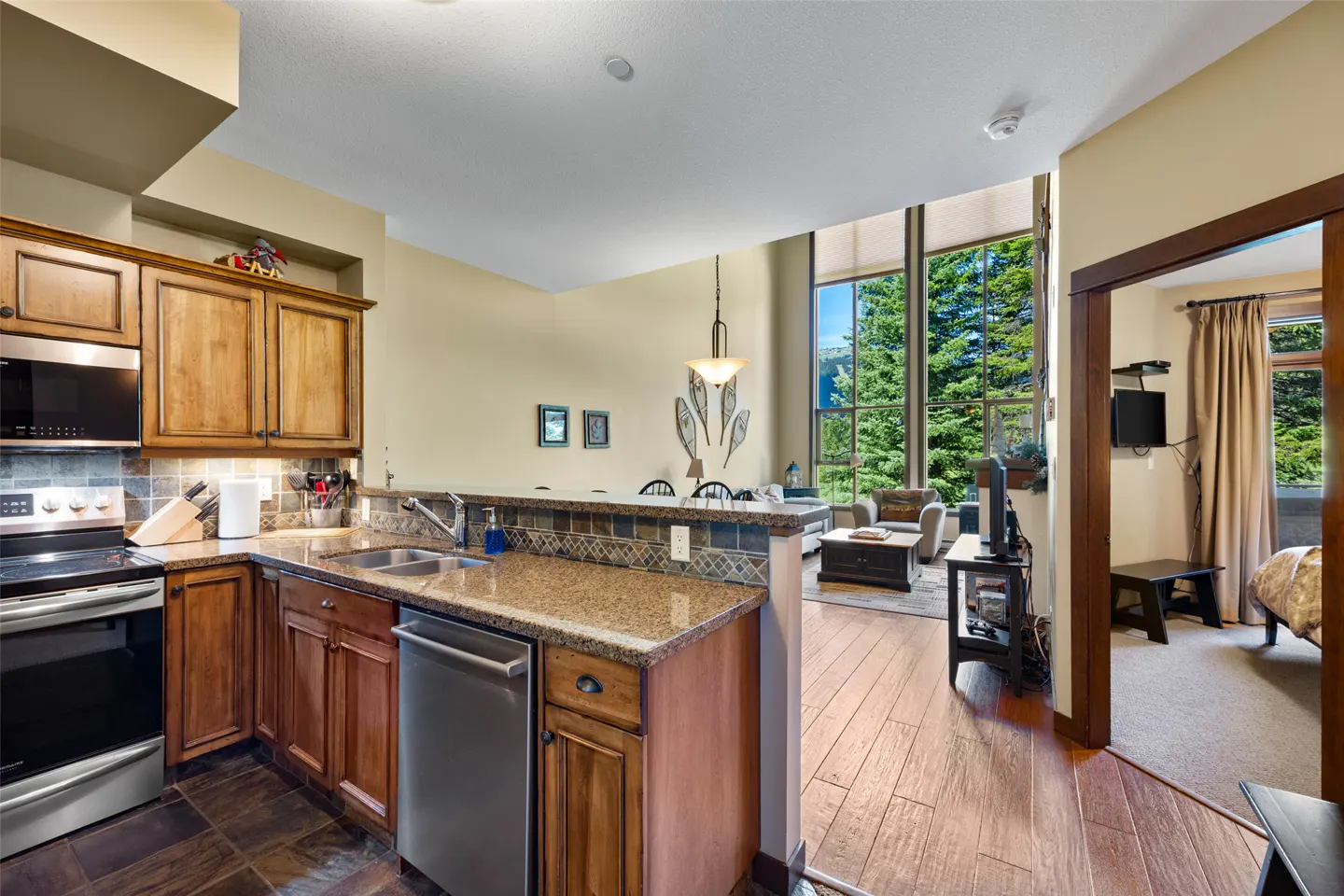 Interior view of a condo with wood cabinets, stainless steel appliances, and a living room with large windows showing trees.