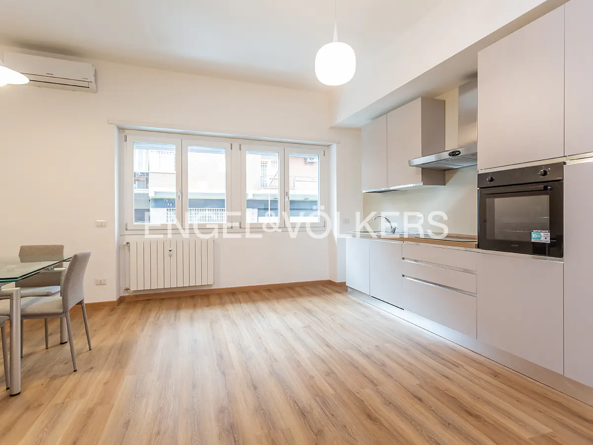 Bright, modern apartment with light wood floors, white walls, and a sleek kitchen. A glass table and chairs sit near a window.