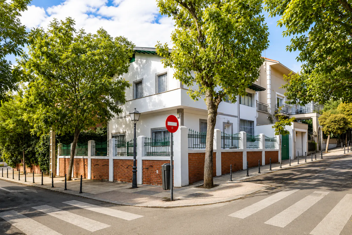 Two-story white house with a brick and white fence, green trees, and a crosswalk in front.