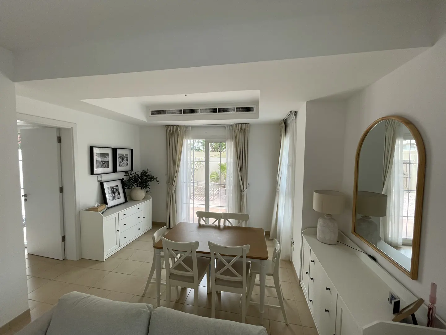 Bright dining room with a wooden table, white chairs, and cabinets. A mirror hangs on the wall. Curtains frame the window.