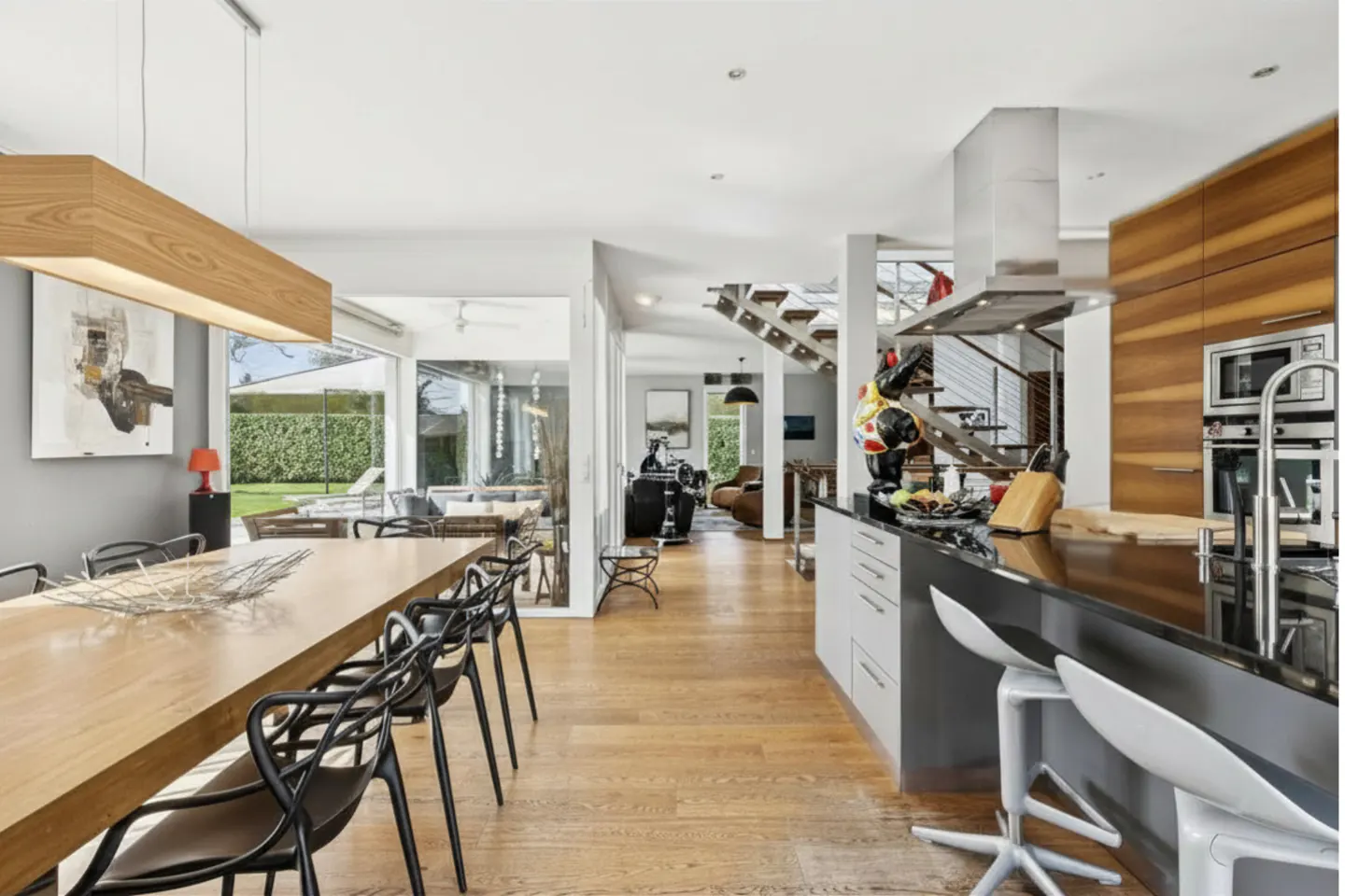 Open-concept home interior with a long wood table, black chairs, and a kitchen island with white stools. A staircase is visible in the background.