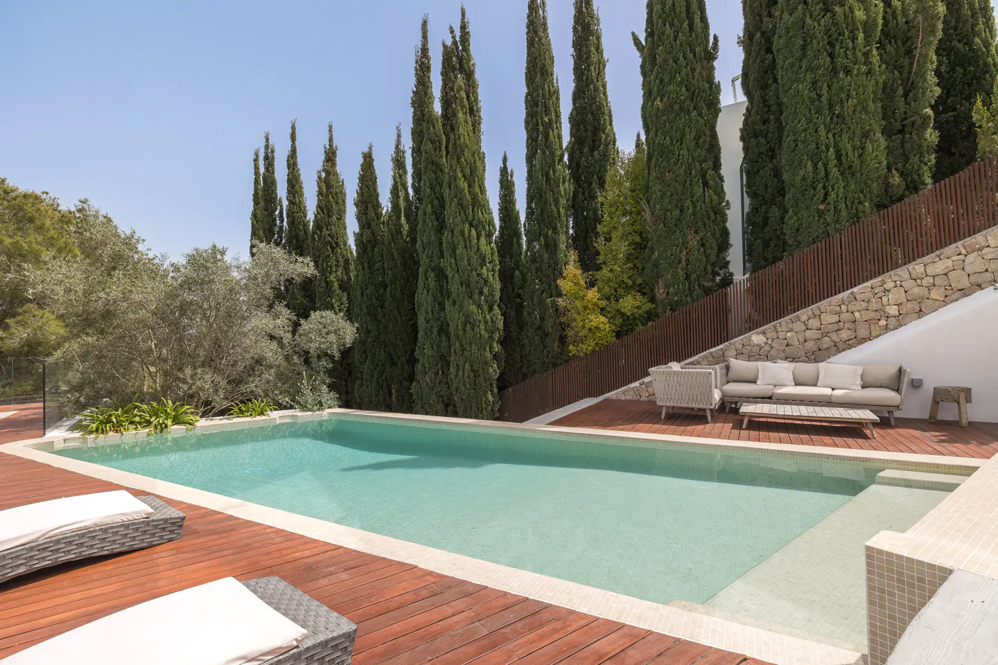 Outdoor pool with wooden deck, lounge chairs, and seating area. Tall green trees and a stone wall provide privacy.