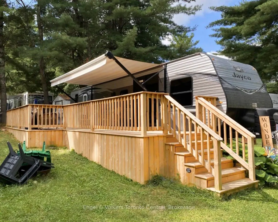 A Jayco RV with a wooden deck and awning sits on a grassy lot surrounded by trees.