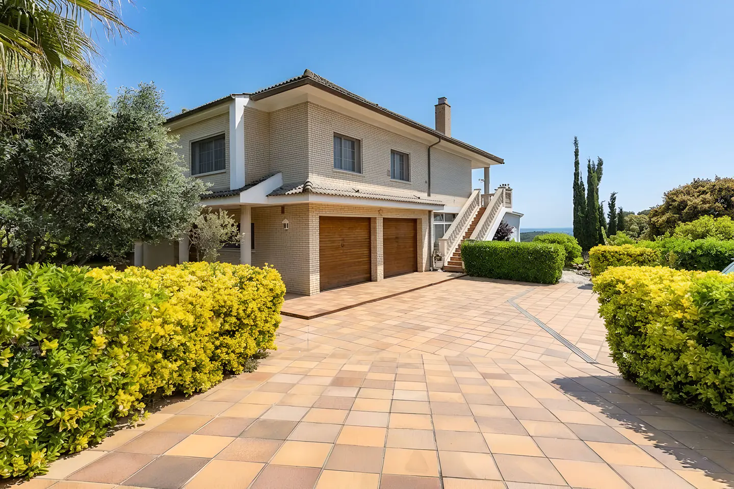 Two-story beige brick house with a two-car garage and a tiled driveway on a sunny day.
