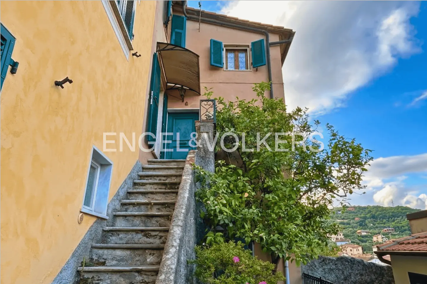 Stone stairs lead to a teal door on a peach-colored building with green shutters and a lush tree against a blue sky.