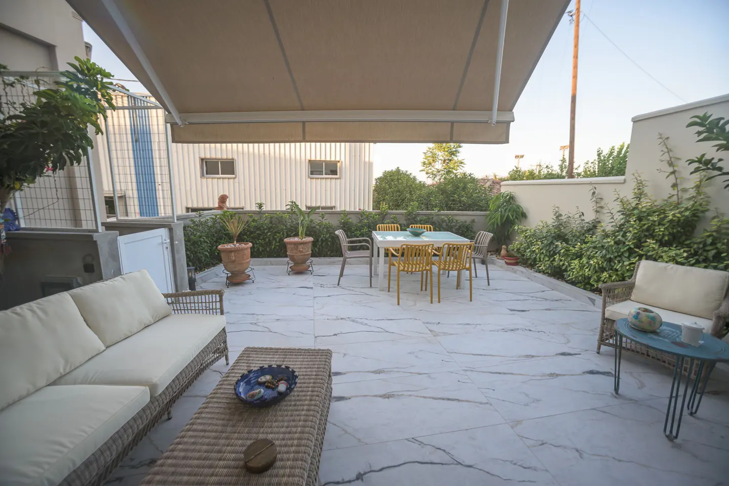 A patio with a white marble floor, a beige awning, and outdoor furniture. A wicker sofa and coffee table are on the left, and a table with yellow chairs is in the center.