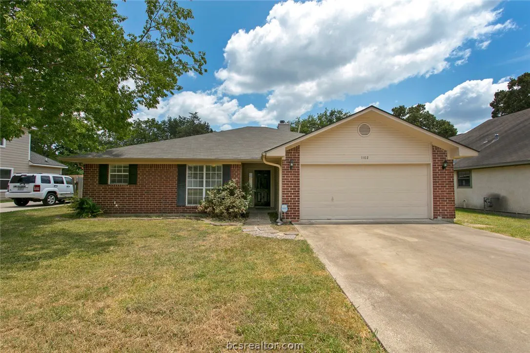 A single-story brick house with a beige garage door and a green lawn under a blue, cloudy sky.