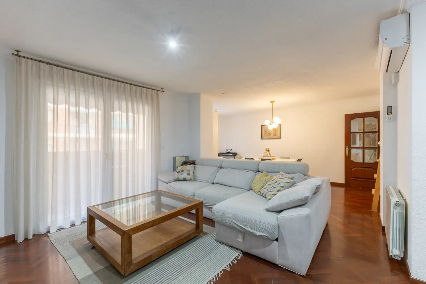 Living room with a gray sectional sofa, glass-topped coffee table on a striped rug, and sheer white curtains.