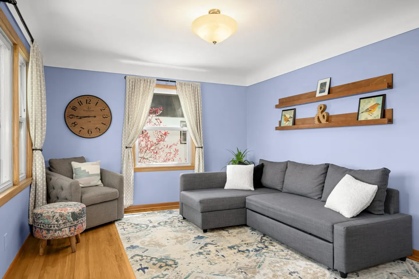 Living room with light blue walls, gray sectional sofa, armchair, and wood shelves with art. A large clock hangs on the wall.