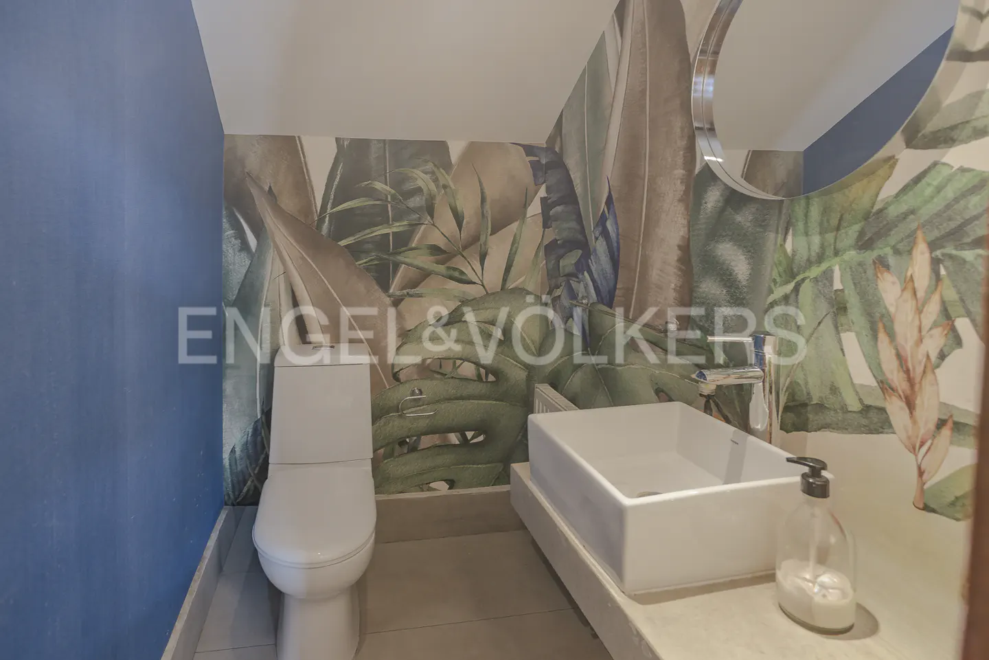 A bathroom with a white toilet and sink, tropical leaf wallpaper, and a blue accent wall.