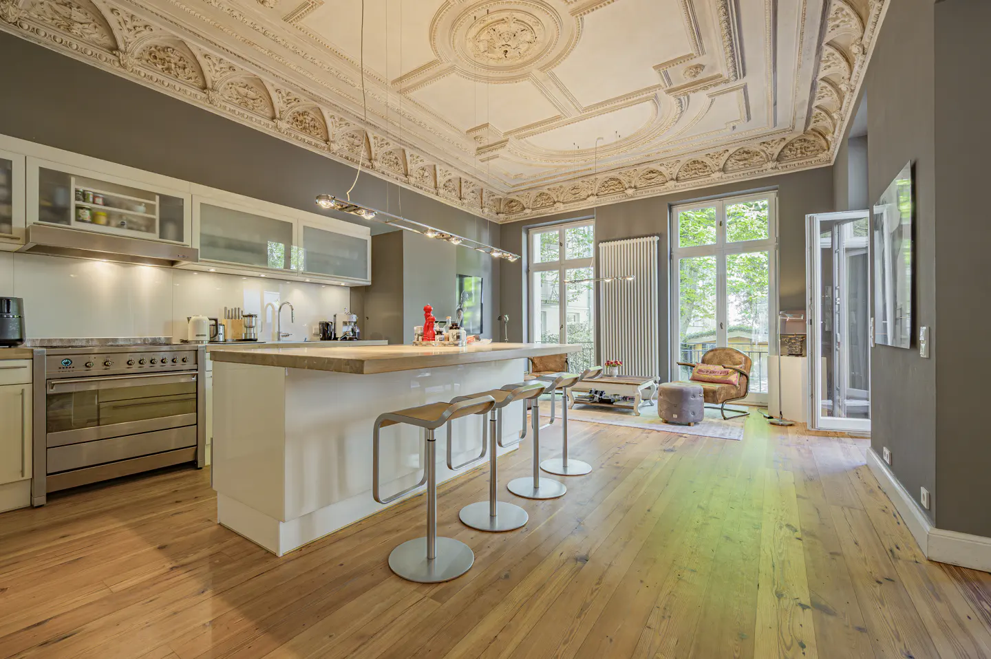 Open-concept kitchen and living room with wood floors, white cabinets, and ornate ceiling. Four stools line the kitchen island.