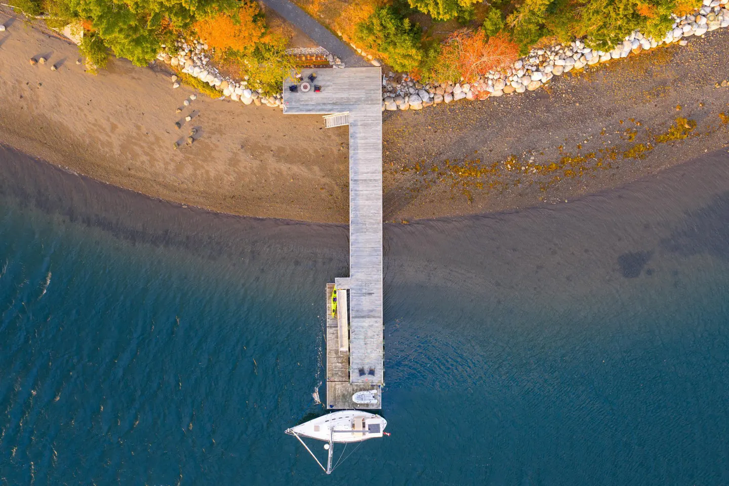 Aerial view of a wooden dock extending into blue water, with a sailboat moored at the end. Autumn foliage lines the sandy shore.