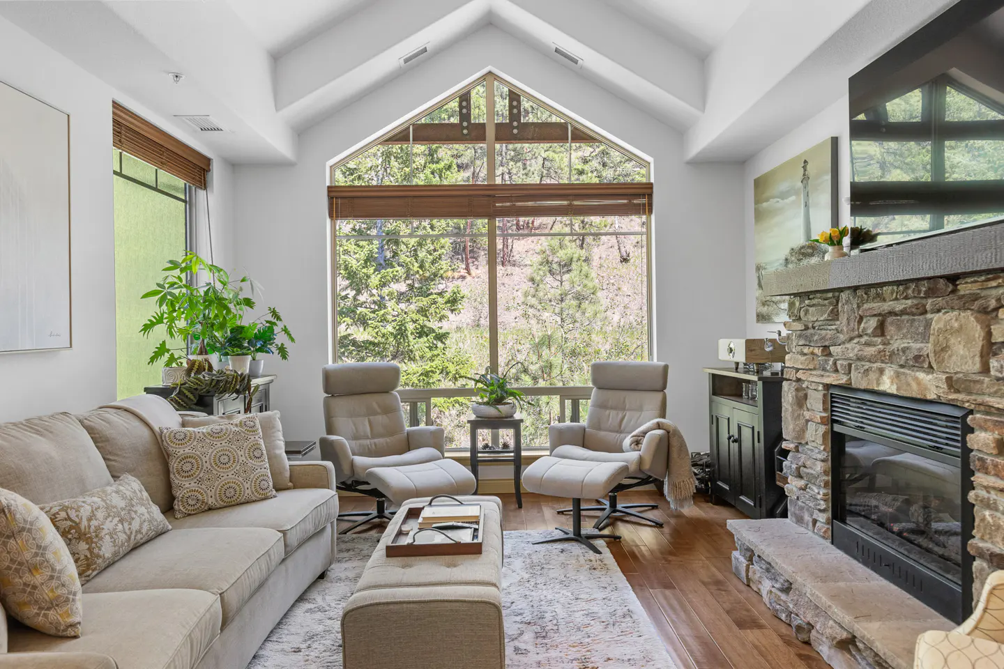 Living room with a beige sofa, two chairs, and a stone fireplace. A large window shows a view of trees.