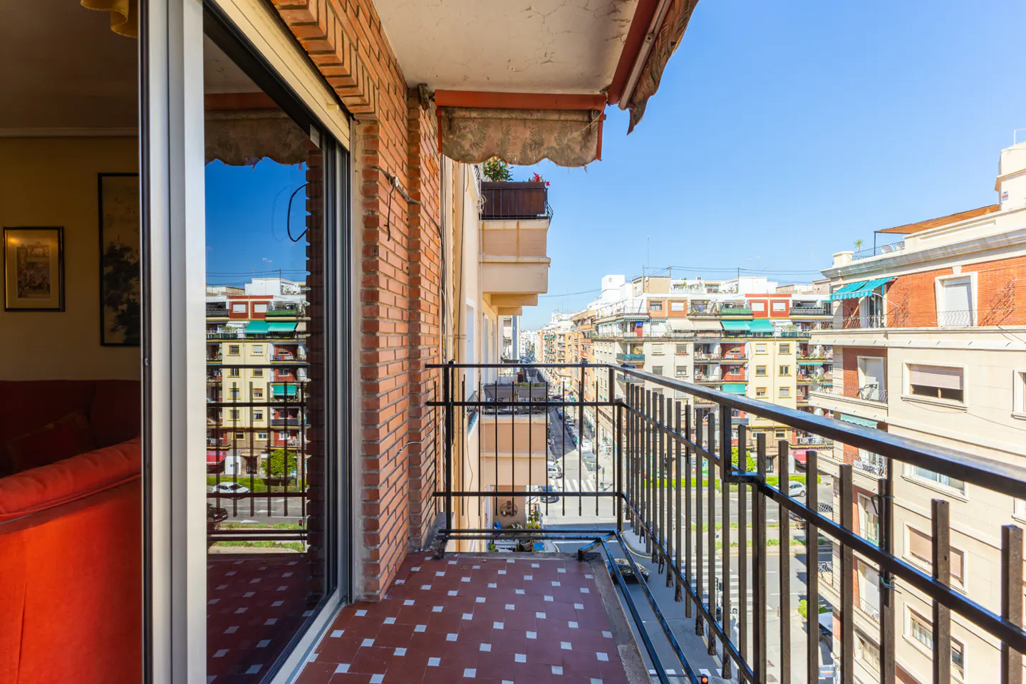 Balcony view with red tile floor and black railing overlooking city buildings on a sunny day.